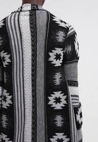 Černobílý vzorovaný kardigan s geometrickými vzory, texturovanou tkaninou a otevřeným předním dílem s dlouhými rukávy.