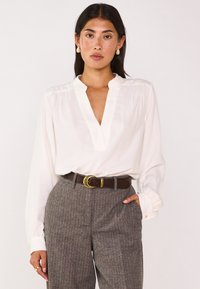 Blusa bianca a maniche lunghe con scollatura a V e polsini con bottoni, abbinata a pantaloni grigi a righe sottili e una cintura marrone con fibbia dorata.