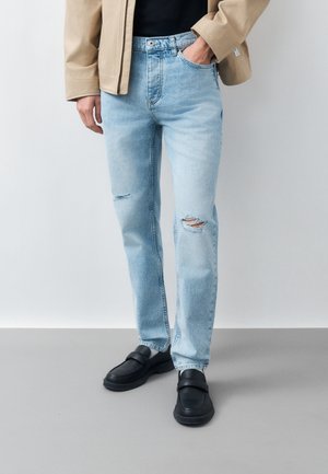 Persoană îmbrăcată în blugi albaștri deschiși rupți, pantofi negri, cămașă neagră și jachetă bej, cu o mână în buzunar, stând pe podea gri.