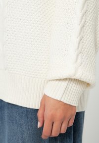 Pulover alb din tricot, cu un corp texturat și manșete cu nervuri, având un model de tricot în cablu pe mânecă pentru un plus de detaliu.