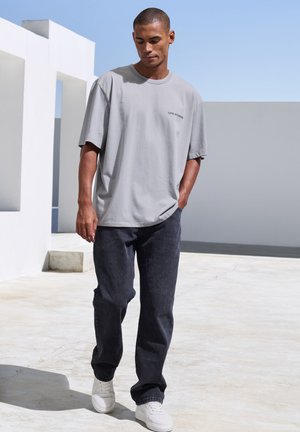Gray oversized t-shirt with logo, dark denim jeans, and white sneakers. Smooth fabric, relaxed fit, and minimalistic design in a vibrant outdoor setting.