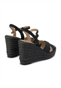 Sandales compensées noires avec une plateforme tressée et texturée. Présente des brides croisées avec de petites clous, une bride réglable autour de la cheville et un design à bout ouvert.