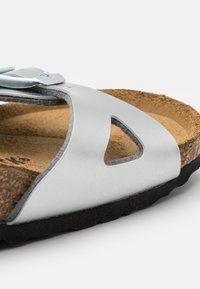 Stříbrná metalická sandál s korkovou stélkou, vyznačující se výřezovým designem, nastavitelným páskem s metalickou sponou a texturovanou černou podrážkou.