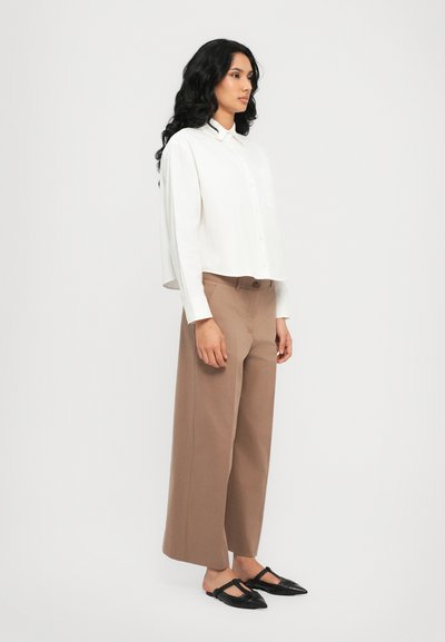 Chemise blanche à boutons raccourcie, pantalon beige à jambes larges et ballerines noires ouvertes. Elle présente des lignes simples et une texture lisse.