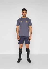 Pelēka sportiska T-krekla ar zelta logotipu, tumši zili šorti, melnas zeķes un melni sporta apavi ar naglām. Vienkāršs dizains ar kontrastējošām krāsām un sportisku pieguli.