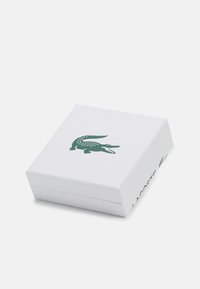 Boîte cadeau blanche avec une finition texturée, arborant un logo crocodile vert sur le dessus et "LACOSTE" en vert sur le côté. Design carré et simple.
