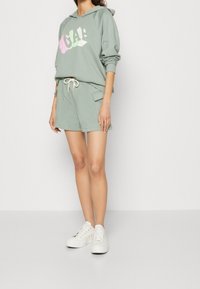 Sweat à capuche vert clair avec le logo "GAP" en blanc et rose, assorti à un short assorti et des baskets blanches. Matière coton, coupe décontractée, cordon de serrage ajustable.