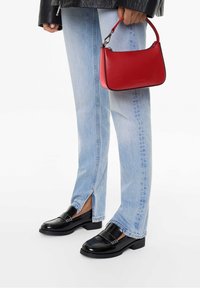 Mocassins en cuir verni noir avec une semelle épaisse, associés à un jean bleu clair avec des fentes latérales. Un petit sac à main en cuir rouge est tenu à la main.