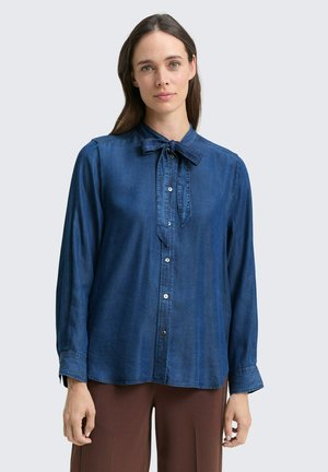 Chemise en chambray bleu à manches longues, avec un devant boutonné et un détail de nœud papillon au cou. Texture douce et coupe décontractée, mise en valeur par des boutons contrastants.