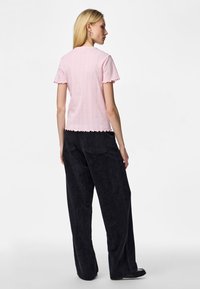 Γυναίκα με πλάτη, φορώντας ανοιχτό ροζ κοντομάνικη μπλούζα με βολάν στις άκρες και μαύρο παντελόνι με φαρδιά μπατζάκια και πίσω τσέπες.