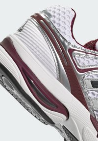 Gros plan du talon d’une chaussure de sport blanche et bordeaux, avec une tige en mesh, une semelle rembourrée et des surcouches synthétiques argentées.