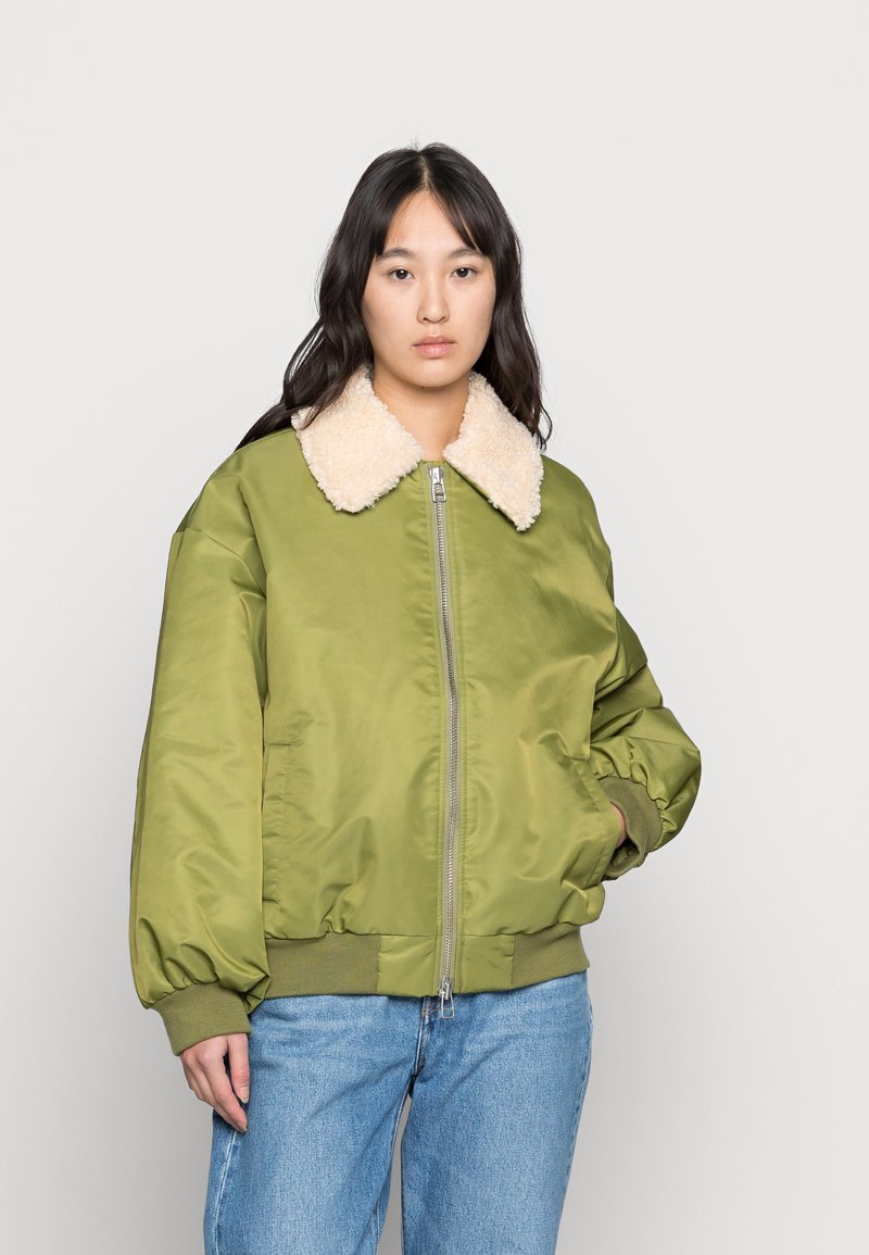 Kobieta ubrana w zieloną kurtkę bomberkę z jasnobeżowym kołnierzem z sherpy i niebieskie dżinsy, stojąca na tle białego tła.