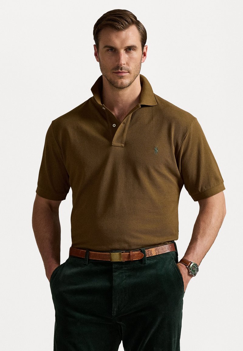 Camisa polo castanha de manga curta e colarinho com três botões, combinada com calças de cotim verde-escuras e um cinto de couro castanho. Emblema icónico exibido.