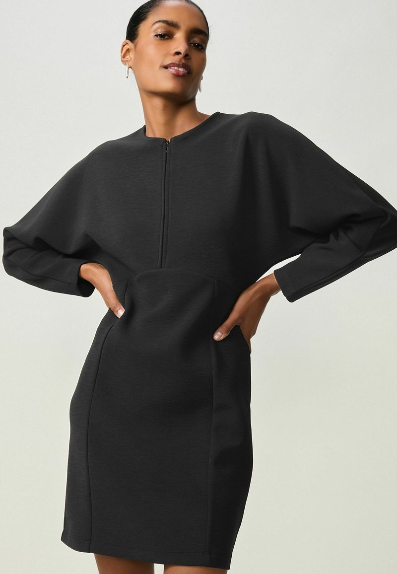 Abito nero con scollatura alta, chiusura con zip davanti e maniche voluminose. Il tessuto è liscio, con una silhouette aderente e tasche laterali.
