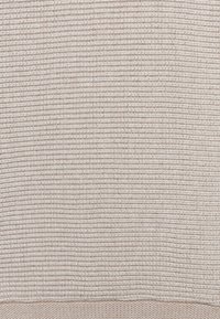 Lys beige strikket stof med et struktureret, ribbet mønster. Materialet virker blødt og åndbart, velegnet til afslappet brug eller hjemmebrug.