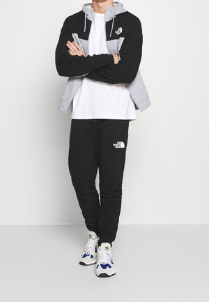 Sort og grå lynlåstrøje over en hvid t-shirt, sammen med sorte joggingbukser med et logo. Hvide sneakers med blå detaljer.