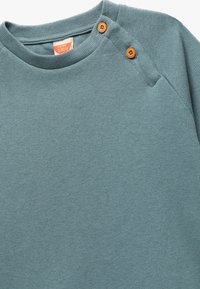 Ljus teal långärmad skjorta gjord av ribbat tyg. Har två träknappar på axeln, rundad halsringning och raglansleeves.