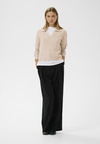 Pulover bej cu dungi, cu decolteu în V și guler, purtat peste o bluză albă, asortat cu pantaloni largi negri și pantofi negri.