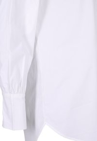 Chemise blanche à manches longues avec poignets froncés et ourlet incurvé sur le côté, présentée sur un fond uni.