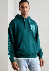Felpa verde scuro realizzata in morbido tessuto, con un logo grafico centrale, cappuccio con cordino regolabile e design floreale sulle maniche.