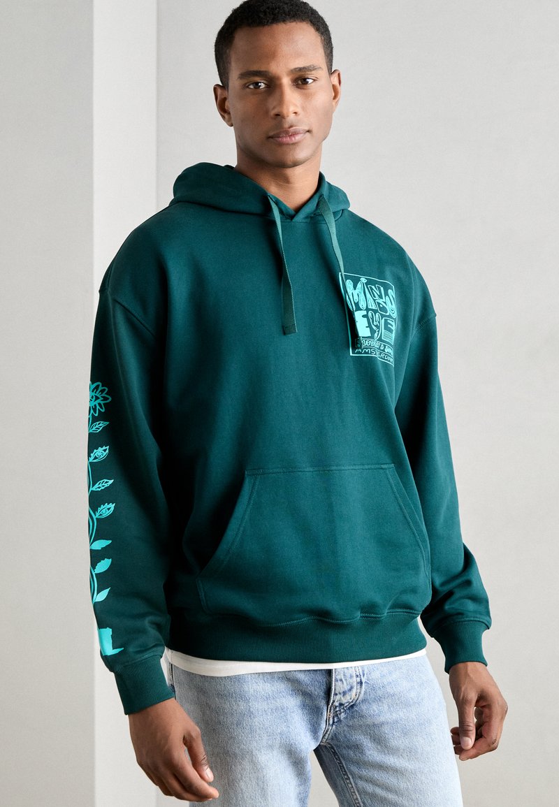 Felpa verde scuro realizzata in morbido tessuto, con un logo grafico centrale, cappuccio con cordino regolabile e design floreale sulle maniche.