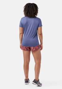 Kortärmad blå skjorta med en slät textur som kombineras med mönstrade röd-orange shorts. Modellen bär grå sneakers. Tydlig vy bakifrån.