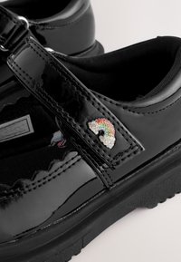 Schwarzer Mary-Jane-Lacklederschuh mit Klettverschluss und kleiner mehrfarbiger Regenbogen-Rhinsteinverzierung am Riemen.
