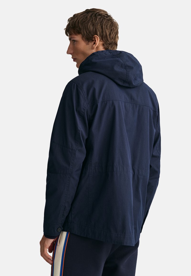 Granatowa kurtka z kapturem o luźnym kroju, charakteryzująca się gładką fakturą, dużym tylnym yoke i zapinanymi na guziki mankietami. Prosty, funkcjonalny design.