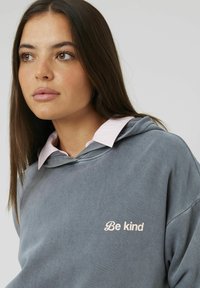 Sweatshirt à capuche gris avec une broderie "Soyez gentil" en rose clair, porté par-dessus une chemise à col rayée rose clair et blanche.