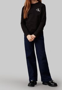 Dlouhý rukáv černá košile s logem a tmavě modré široké kalhoty, doplněné lesklými černými šněrovacími botami. Jednoduchý design bez vzorů.