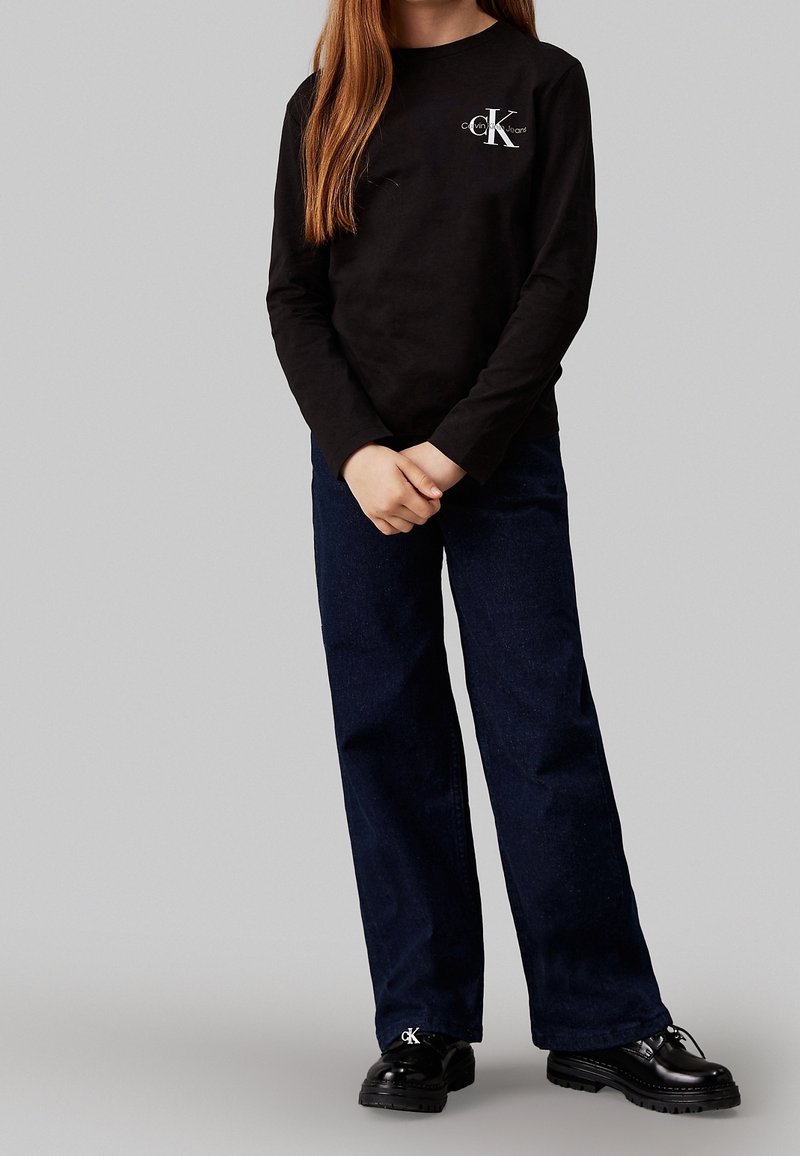 Dlouhý rukáv černá košile s logem a tmavě modré široké kalhoty, doplněné lesklými černými šněrovacími botami. Jednoduchý design bez vzorů.