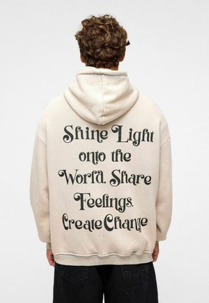 Beige hoodie met een grote zwarte tekstprint op de achterkant die luidt: "Laat Licht op de Wereld Schijnen, Deel Gevoelens, Creëer Verandering." Hoodie heeft een capuchon met trekkoord.
