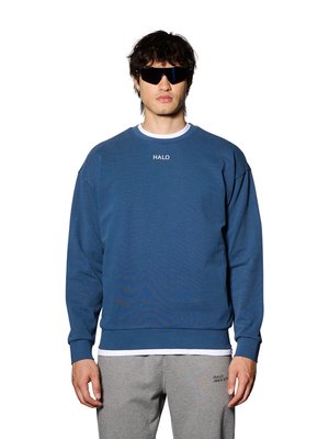 Mann trägt blauen Sweatshirt mit dem Schriftzug "HALO", graue Jogginghose mit kleinem Text und schwarze Wraparound-Sonnenbrille, steht vor weißem Hintergrund.