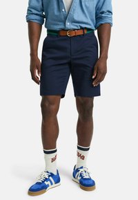 Marineblå shorts, brun belte med grønn stripe, lyseblå skjorte, hvit T-skjorte, blå joggesko, hvite sokker med rødt tekst, avslappet passform.