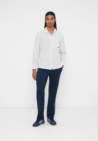 Weißes Langarmhemd mit navyblauen vertikalen Streifen, kombiniert mit navyblauen Hosen mit weißen Seitenstreifen und blauen Sneakers.