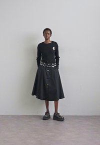 Una persona è in piedi contro uno sfondo neutro, indossando un maglione nero con una toppa ricamata e una gonna nera con una cintura a faccina sorridente.