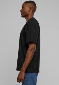 Black cotton t-shirt with short sleeves, relaxed fit, and crew neckline. Side view shows straight cut and minimal detailing. Worn with blue jeans.