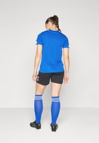 Mujer con el pelo trenzado que lleva una camiseta deportiva azul, pantalones cortos negros, calcetines azules hasta la rodilla y botas negras, de pie de espaldas sobre un fondo liso.