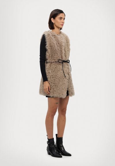 Beige nepleer vest met een textuur aan de oppervlakte, open voorkant en een zwarte strikriem in de taille, gedragen over een zwart shirt met lange mouwen en zwarte shorts.