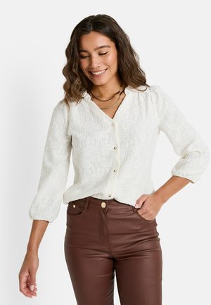 Blouse blanche texturée avec un devant à boutons, des manches trois-quarts et un motif de broderie subtil. Assortie avec un pantalon brun ajusté.