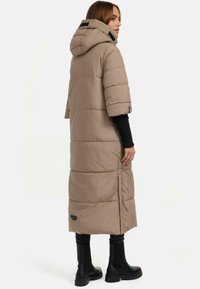 Beige Daunenjacke mit Kapuze, kurzen Ärmeln und einer langen Silhouette. Sie verfügt über horizontale Nähte und ein schwarzes Etikett am Saum.