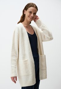 Cardigan bianco e peloso con apertura frontale, due tasche frontali, polsini a costine e una vestibilità rilassata, indossato sopra un top scuro e pantaloni blu navy.