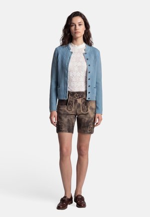 Vrouw draagt een lichtblauwe cardigan met knopen, een witte kanten blouse, bruine geborduurde leren shorts en donkerbruine schoenen, staand tegen een witte achtergrond.