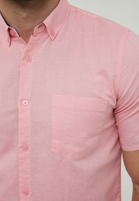 Chemise rose clair à manches courtes en tissu texturé, avec un devant à boutons, un col et une poche appliquée unique sur la gauche.