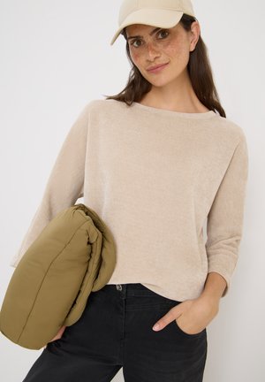 Beiger strukturierter Pullover mit Dreiviertelärmeln, kombiniert mit olivgrüner Steppjacke, über schwarzen Jeans getragen. Cremefarbene Baseballkappe.