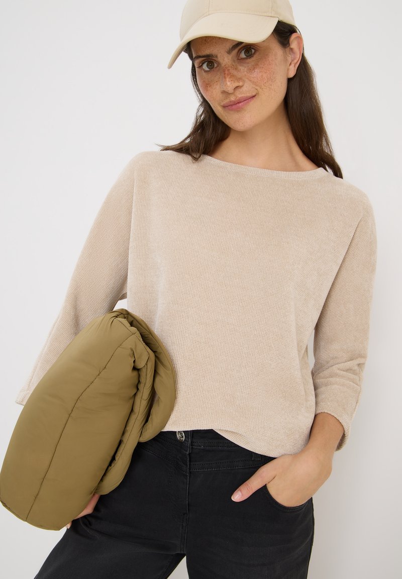 Pull beige texturé à manches trois-quarts, associé à une veste rembourrée verte olive, porté sur un jean noir. Casquette de baseball crème.
