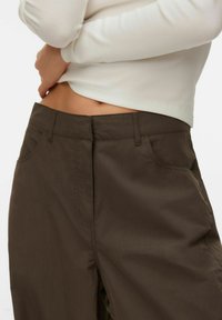 Femme portant un pantalon marron foncé et un haut blanc à manches longues côtelé, légèrement relevé pour dévoiler le bas de l'abdomen.
