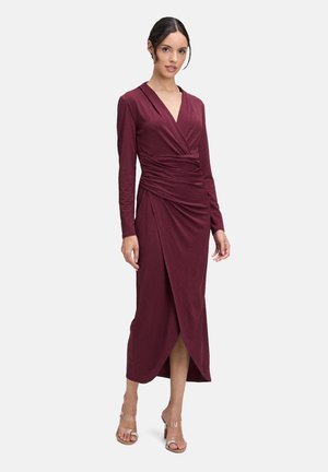 Vrouw in een diep bordeauxrode wikkeljurk met lange mouwen, een gerimpelde taille en een asymmetrische zoom, gecombineerd met doorzichtige hakken.