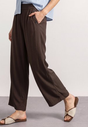 Personne portant un pantalon ample marron foncé avec des boutons latéraux et des sandales plates beiges à bout ouvert, marchant sur un sol gris devant un fond uni.