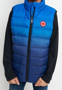 Modrá gradientová péřová vesta s horizontálním prošíváním, vysokým límcem a předním zipem. Obsahuje kulatou červenou nášivku na prsou.